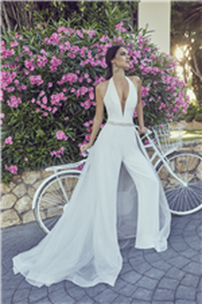 Brunette lady leaning on bike next to pink flowered bush wearing bridal jumpsuit with plunge v neckline and chiffon overskirt