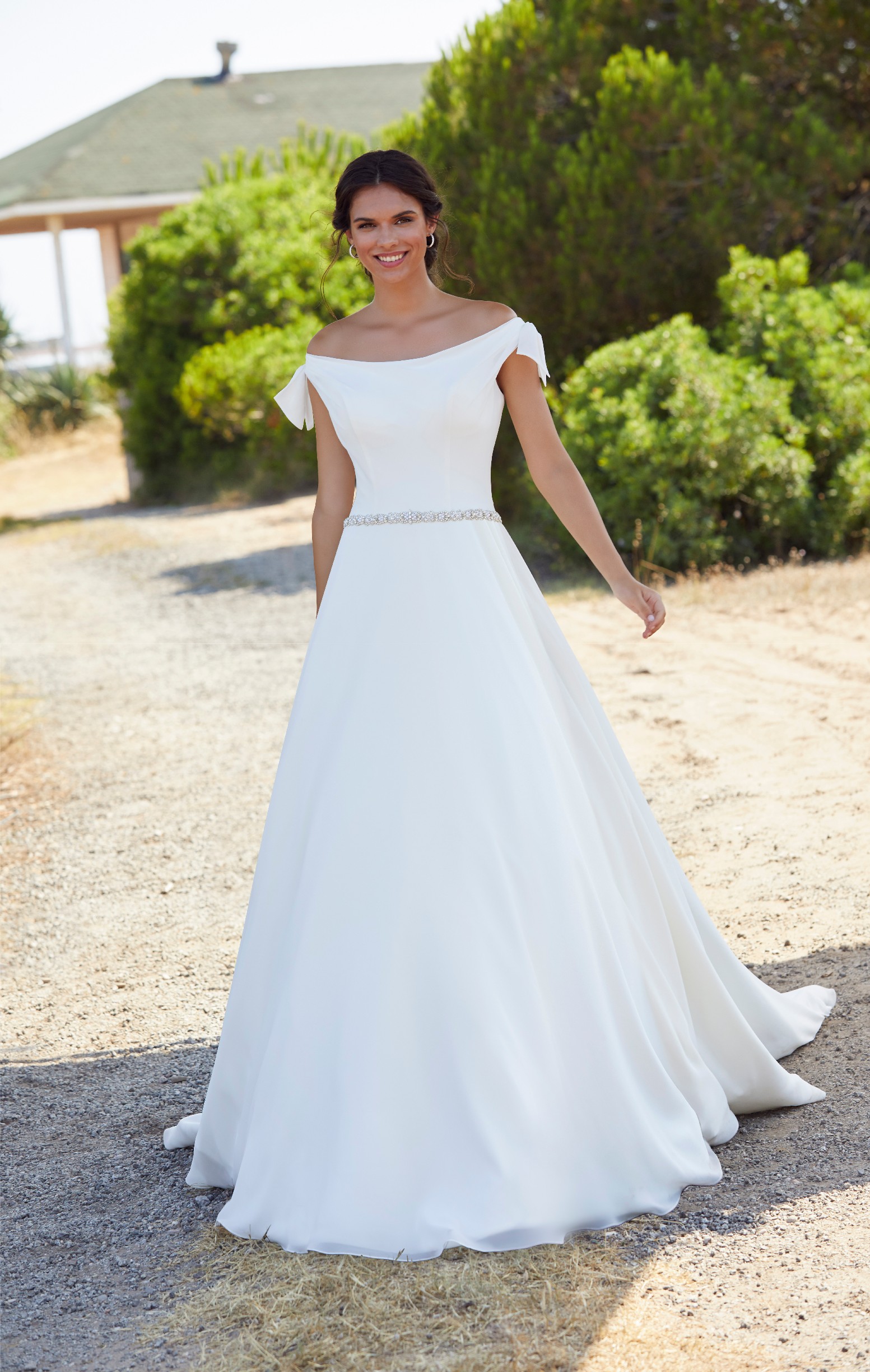 Brunette bride stood on grass by green bushes in Ronald Joyce 18407, a plain ivory A-line wedding dress with off the shoulder cap sleeves and a delicate sparkle belt