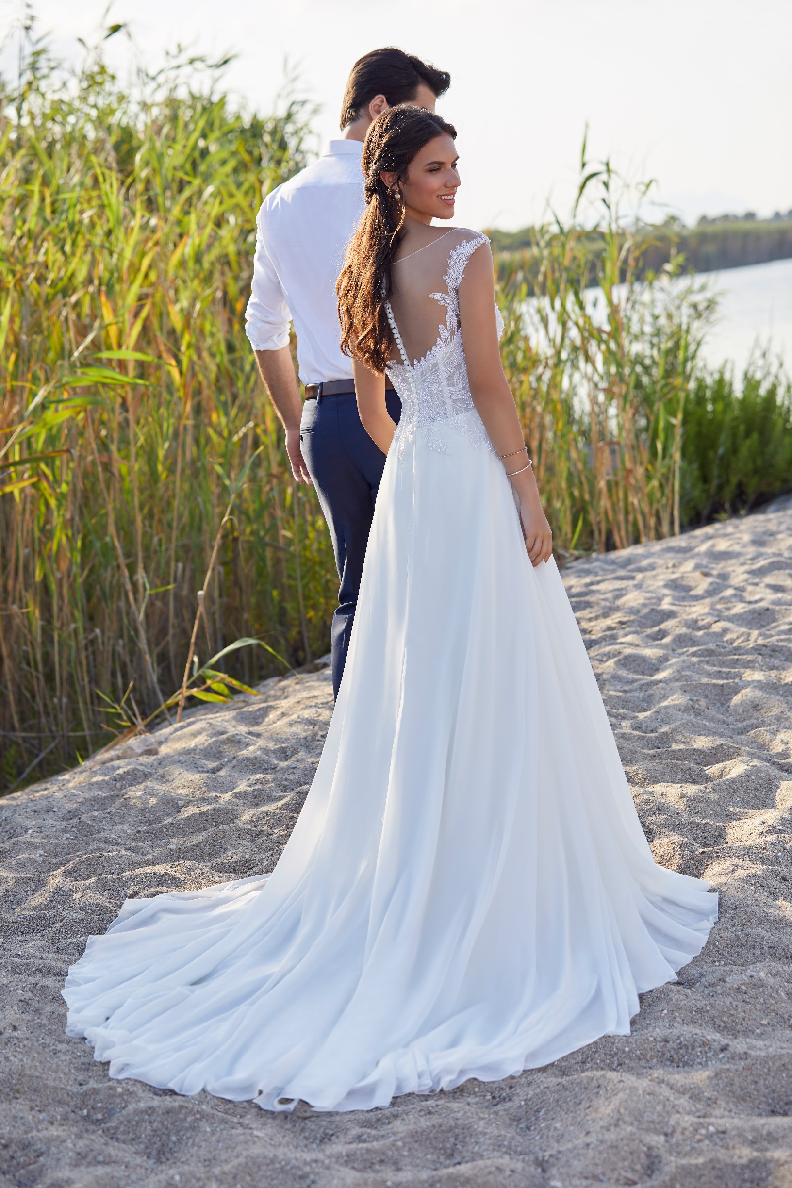 Thumbnail photo of the back of bride and groom models stood by tall reeds on a beach. Bride wears Ronald Joyce 18413, a white A-line wedding dress with an illusion necklace back