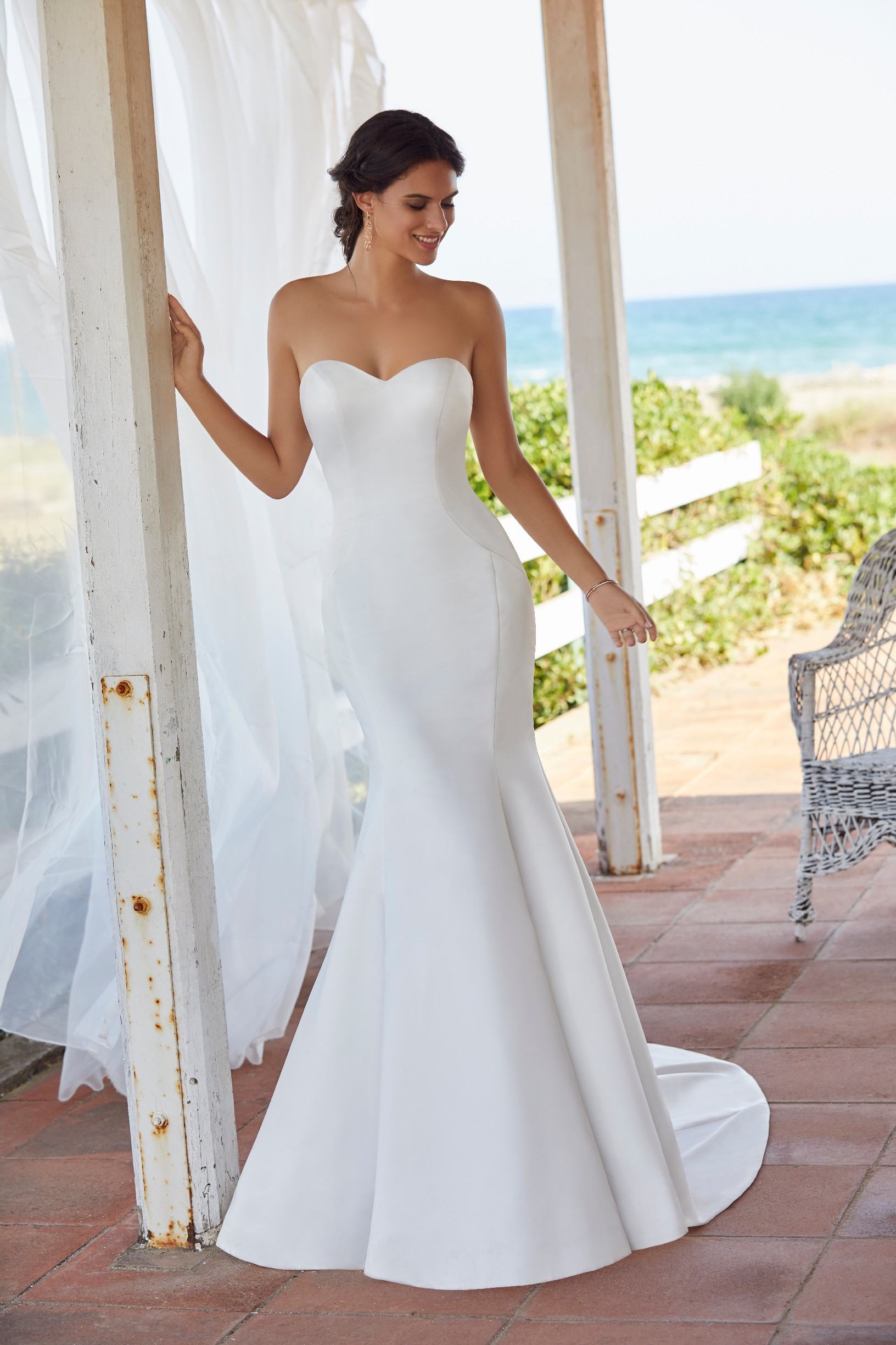 Model stood on a terracotta terrace by the sea in Ronald Joyce 18417, a plain strapless wedding dress with bow back detail suited to a destination wedding. 