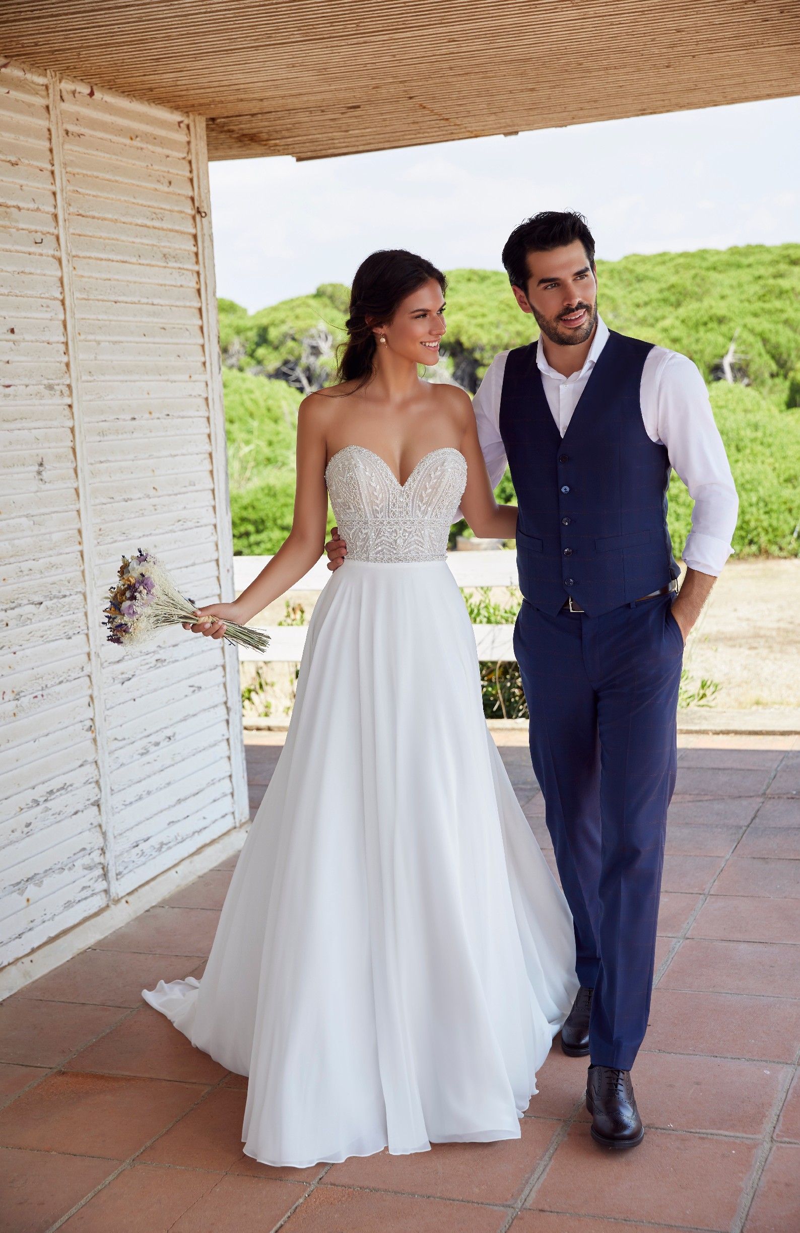 Bride and groom stood arm in arm on a terrace in front of Italian greenery. Model wears Ronald Joyce 18419, a strapless wedding dress with a beaded sweetheart bodice and plain skirt