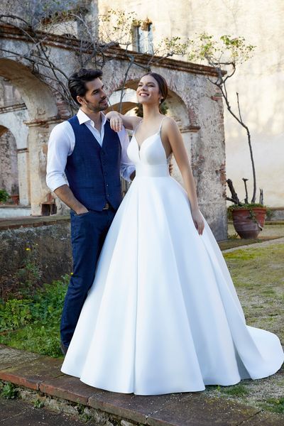 Woman standing in garden wearing ballgown wedding dress with spaghetti straps and leaning on man