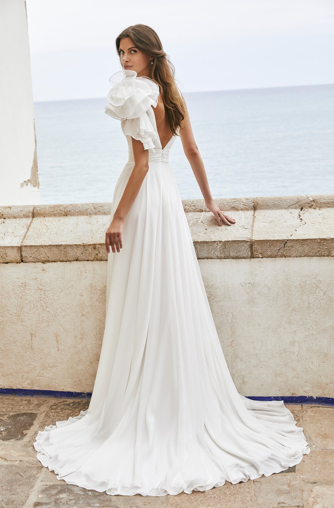 Back profile of model in Ronald Joyce destination wedding dress style 18602, a floaty chiffon A-line dress with a leg split, ruched waistband, floral shoulder detail, and open back.