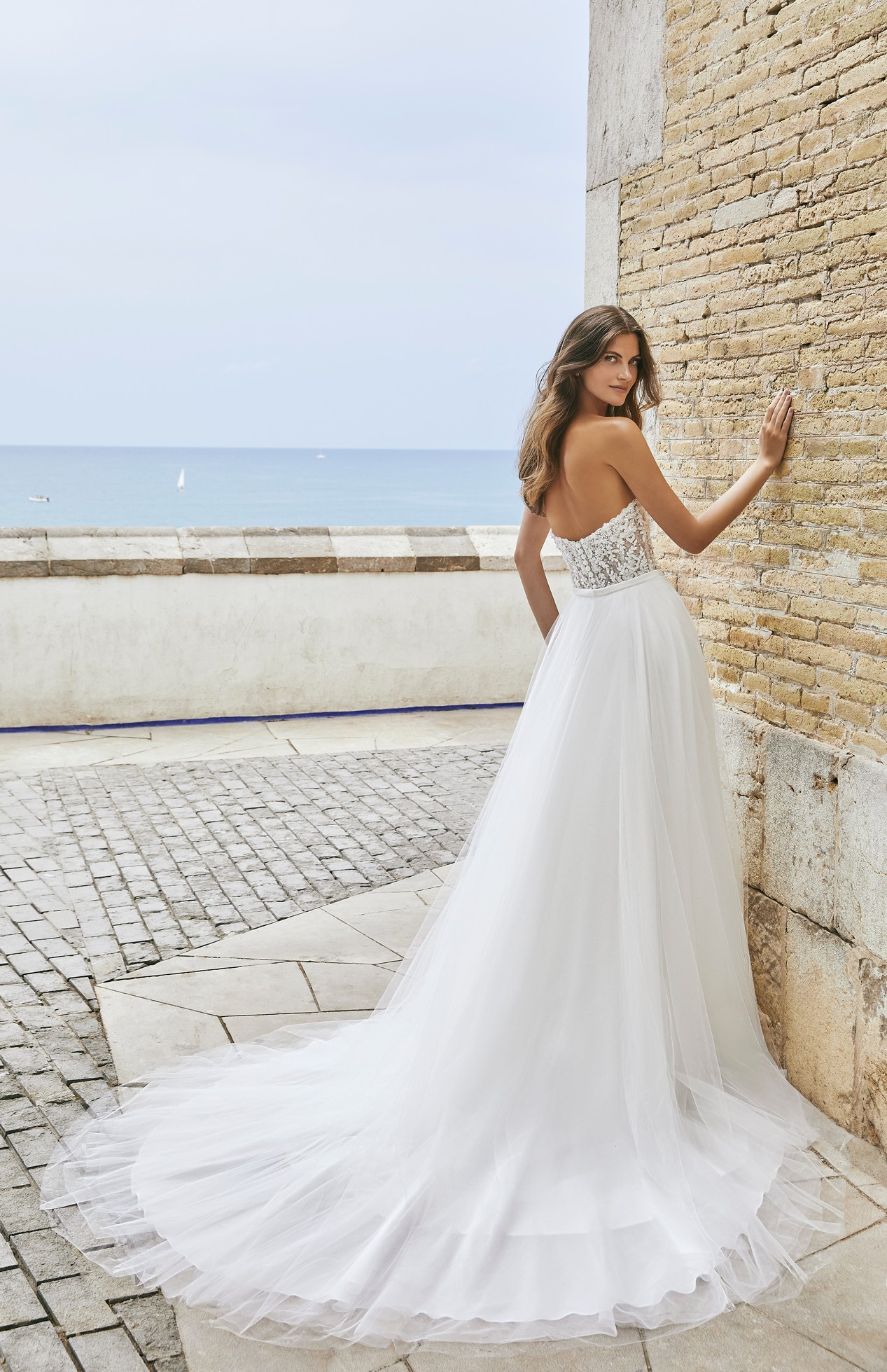 Back profile of model wearing Ronald Joyce Victoria Jane destination wedding dress style 18604 with matching overskirt. Model stands against a wall by the sea.  