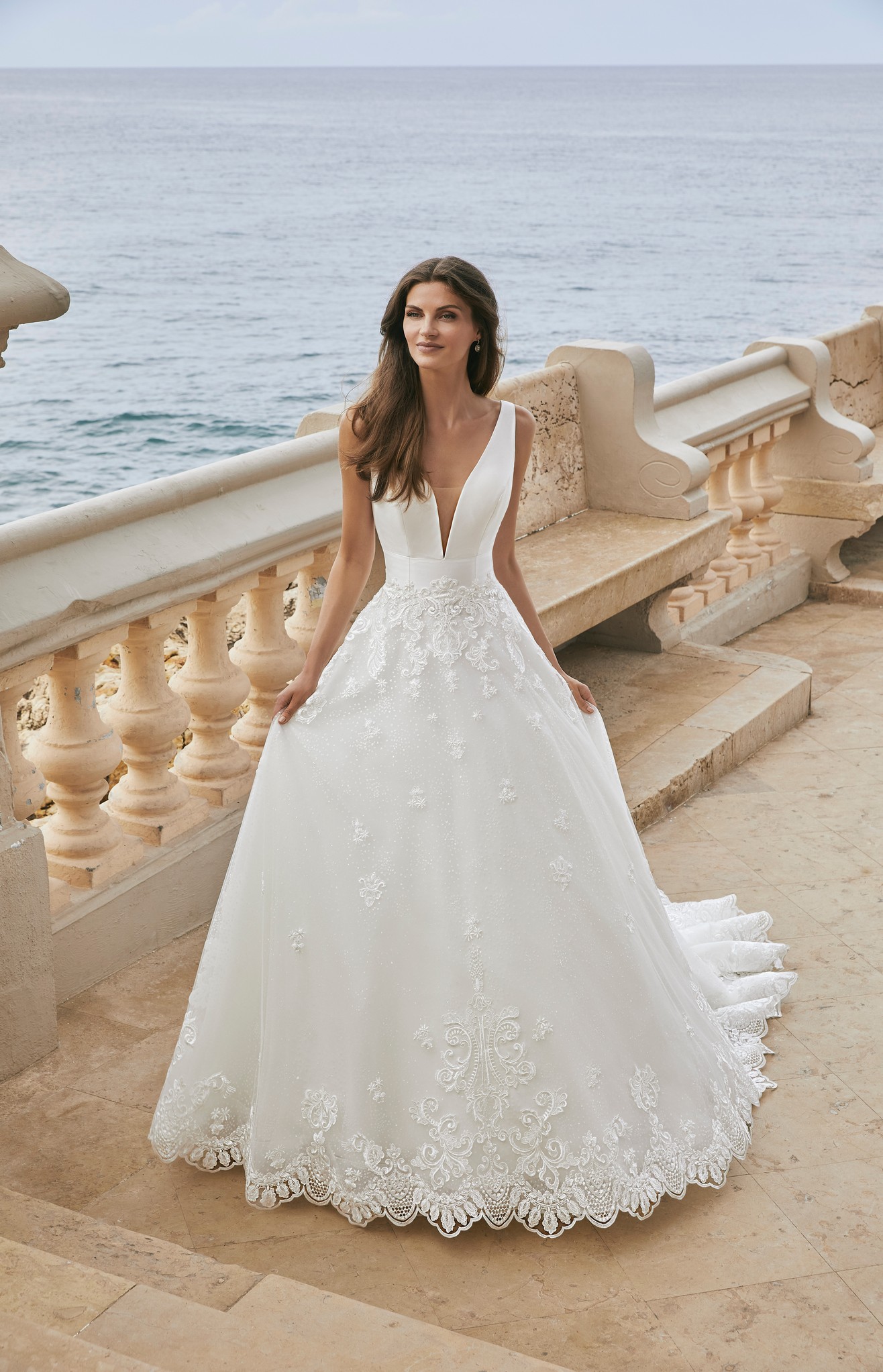 Model stood by the sea in Ronald Joyce style 18612, a ballgown wedding dress with a plunging illusion v-neckline, open back and glitter tulle and lace applique skirt. 