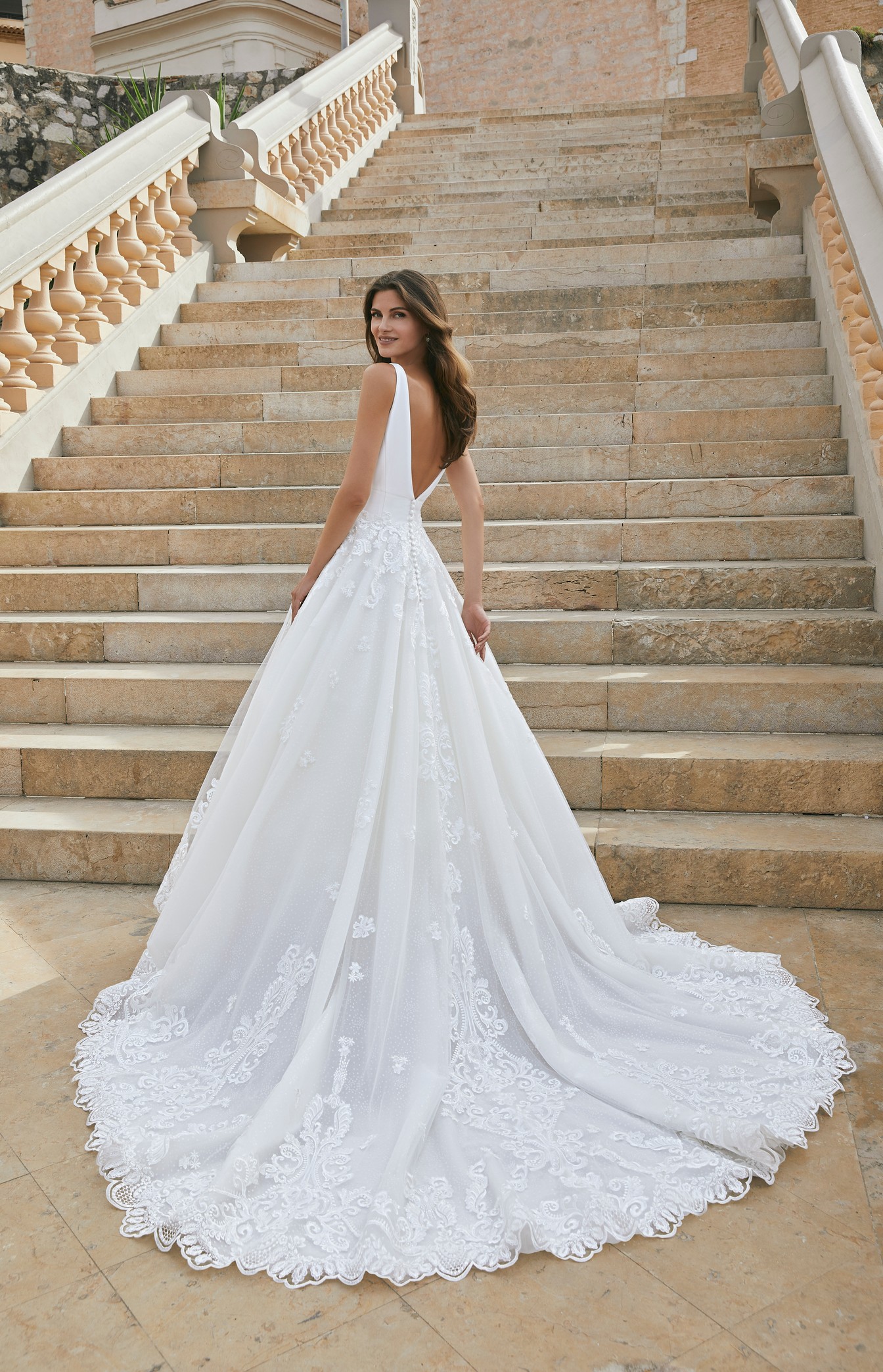 Back of model stood by outside steps in Ronald Joyce style 18612, a ballgown wedding dress with a plunging illusion v-neckline, open back and straps.