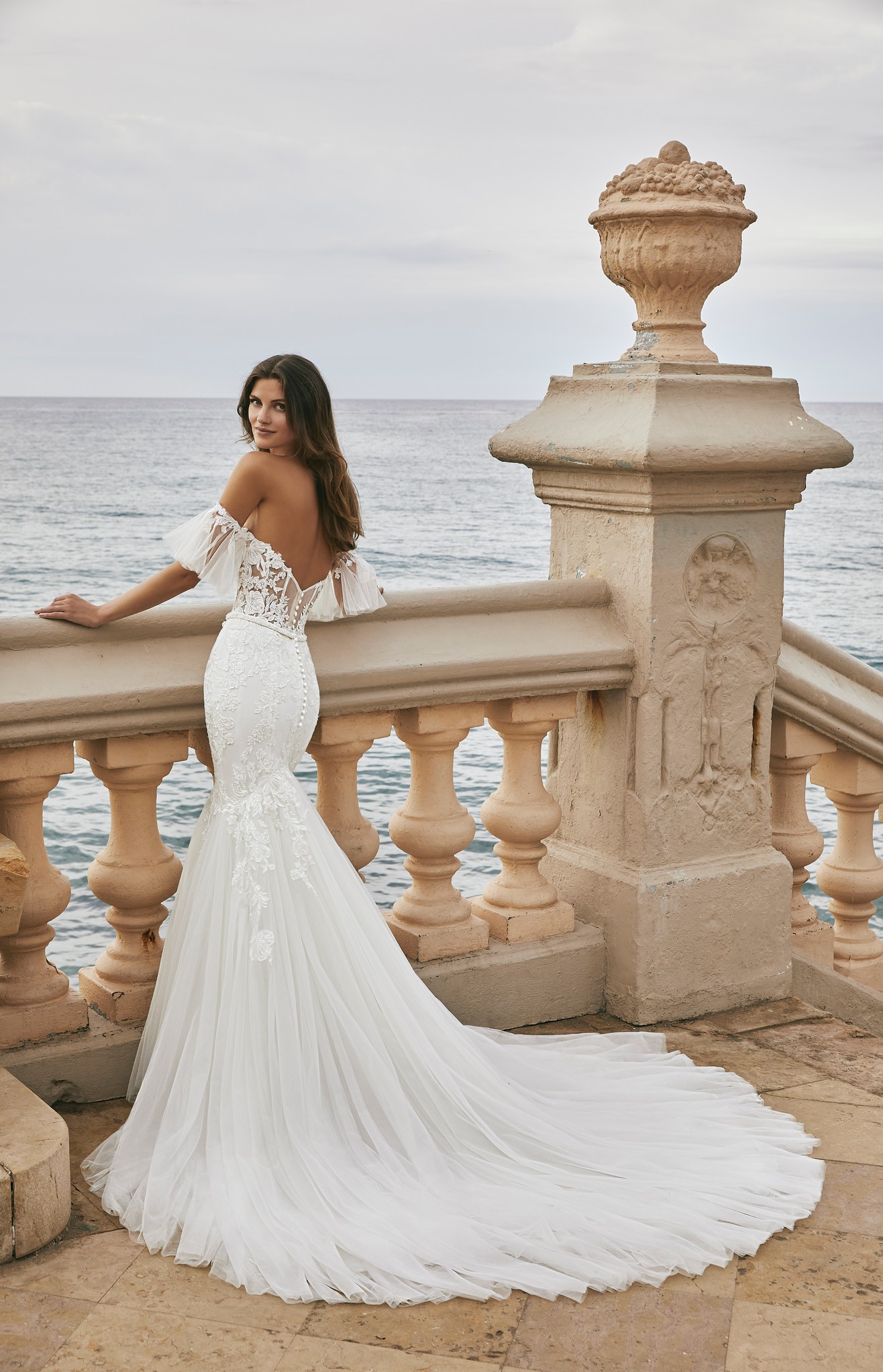 Back of model stood by the sea in Ronald Joyce style 18615, a strapless fit and flare wedding dress with a tulle skirt, illusion lingerie-inspired bodice and detachable sleeves