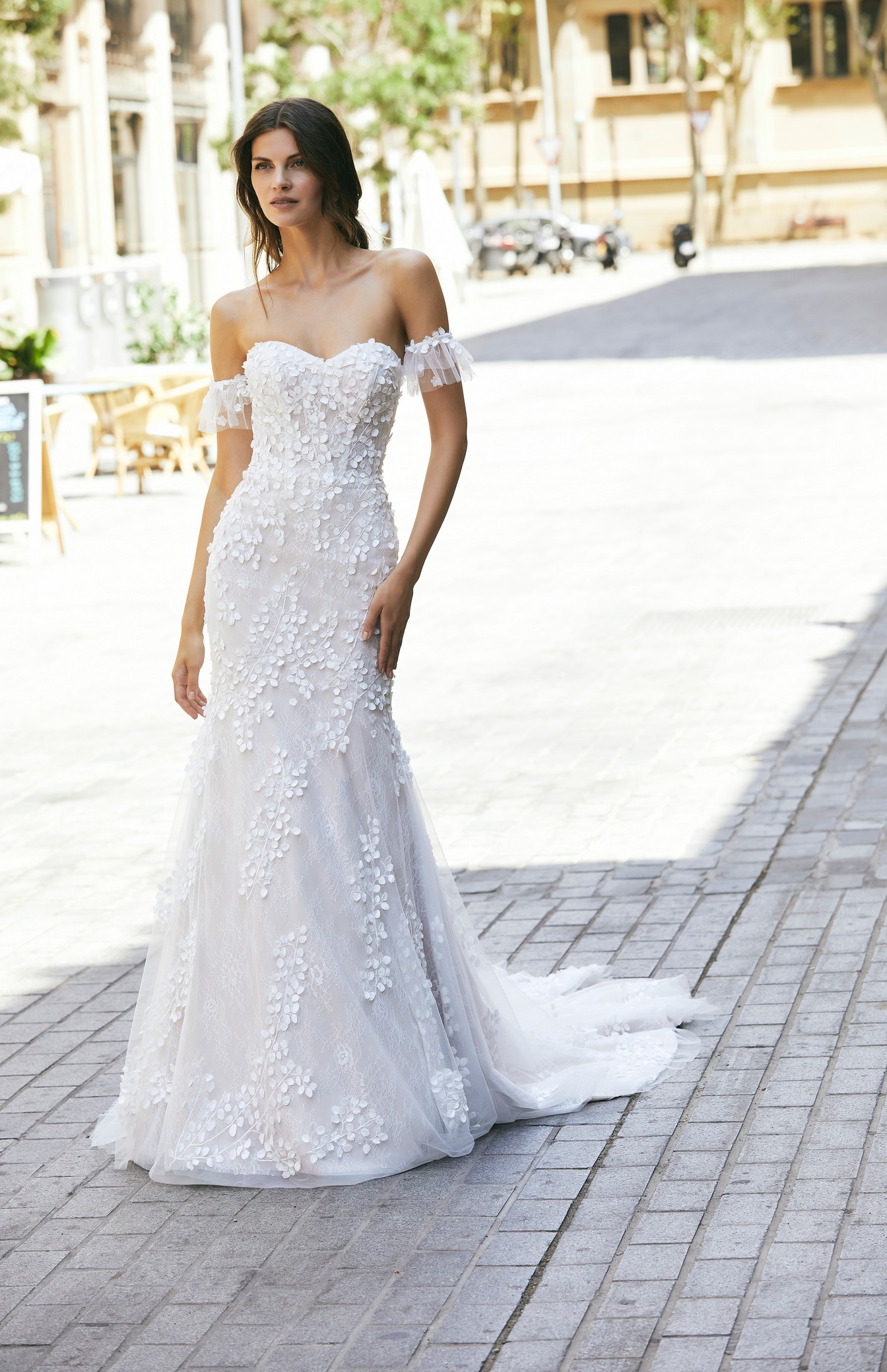 A brunette model stood in an empty sunny street in Ronald Joyce 18616, a strapless floral applique wedding dress style with a subtle sweetheart neckline and detachable cap sleeves.