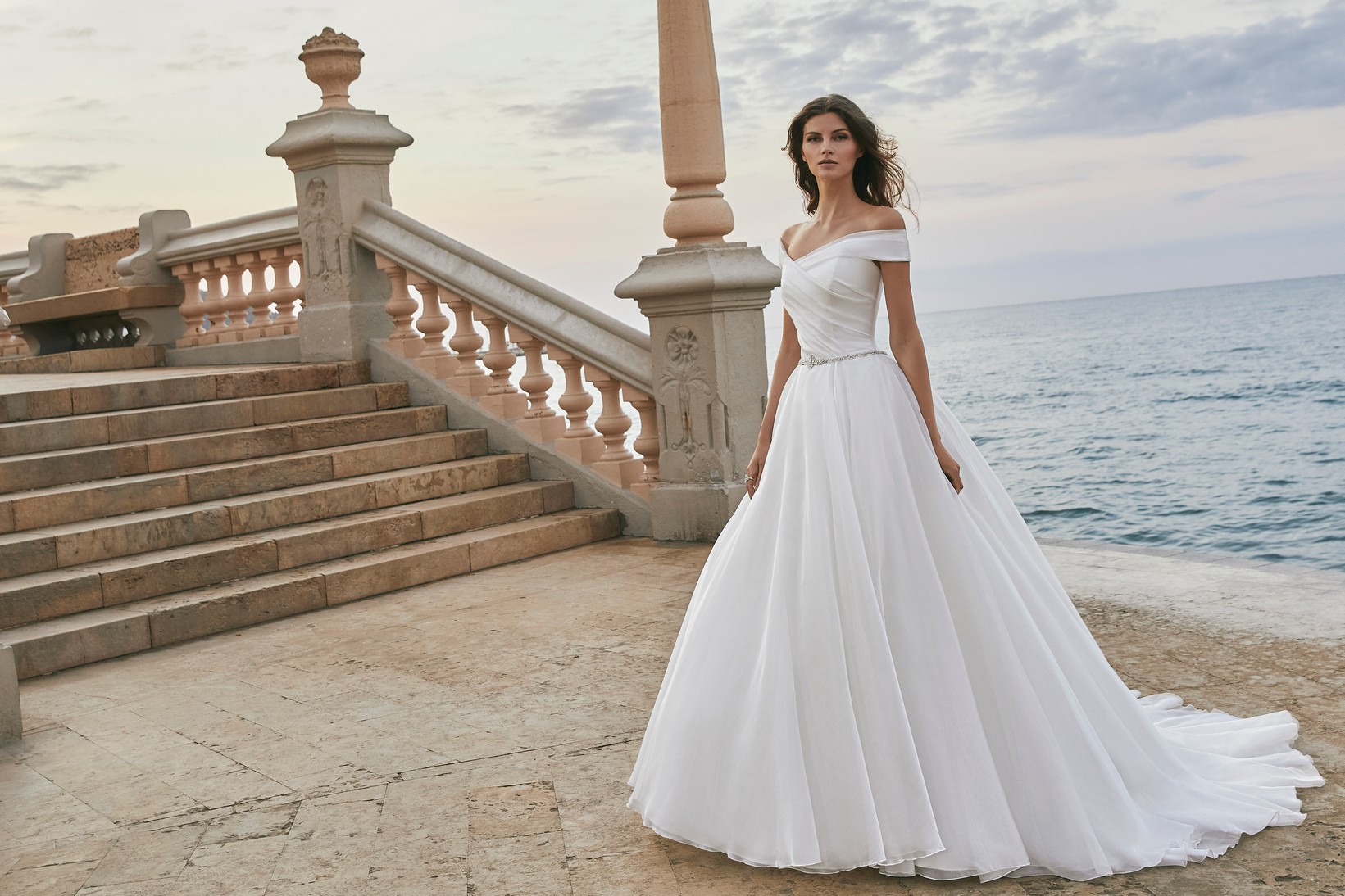 Model wearing Ronald Joyce style 18619, a plain off the shoulder ballgown wedding dress with a delicate diamante belt and integrated cathedral train.