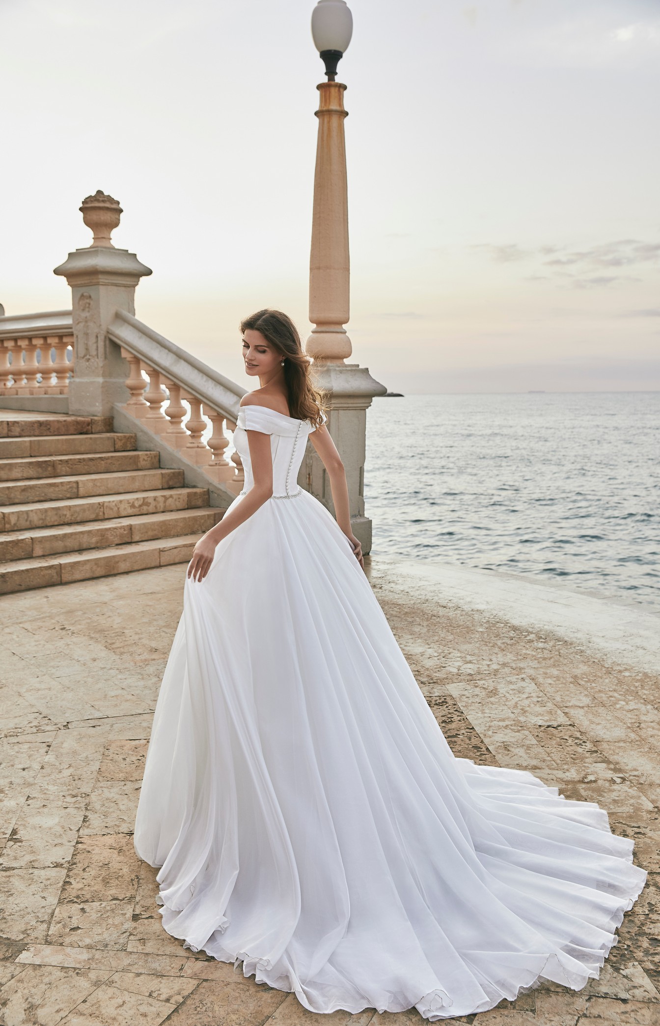 Back of a brunette model stood by steps by the sea in Ronald Joyce 18619, an off-the-shoulder fairytale ballgown wedding dress with a delicate diamante belt and closed button back