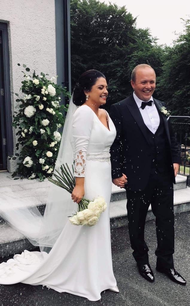 Real bride Emma stood outside holding hands with husband Brian in Ronald Joyce Tai 18202, a fitted sheath wedding dress with long lace cuff sleeves, an embellished waist and v neckline.