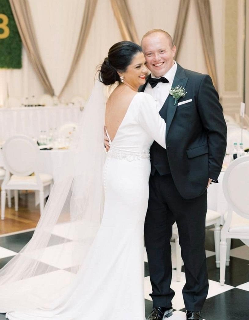 Close up of real bride Emma stood smiling and hugging husband Brian. Her back is to the camera, showing a floor-length tulle veil and the v-shape open back of her white Ronald Joyce 18202 wedding dress