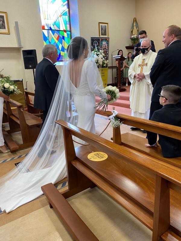 Real bride Emma approaches her husband at the altar. She holds a bunch of white roses and wears Ronald Joyce 18202 Tai wedding dress with a floor-length tulle veil over her face.