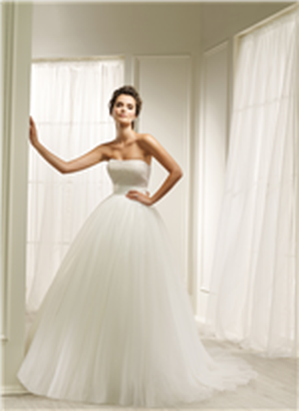 Brunette lady standing in front of white house background wearing strapless, beaded ballgown wedding dress