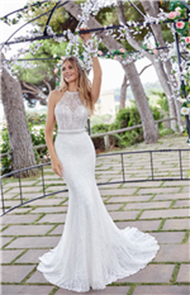 Blonde woman standing under floral decorated arch wearing beaded bodice wedding dress with high neckline and embellished waist belt