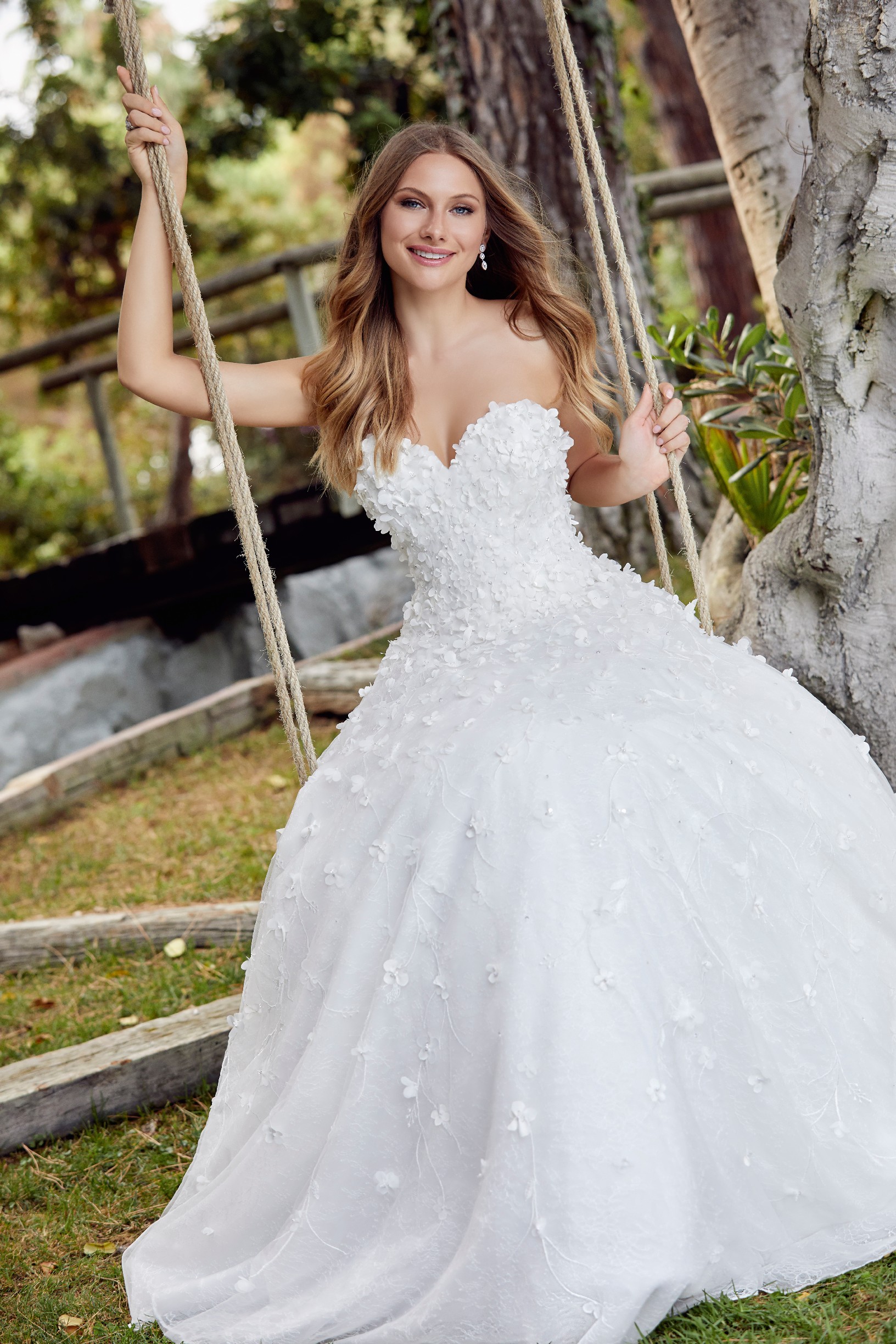 Blonde woman in the garden on tree swing wearing strapless ballgown wedding dress with 3D floral detail 