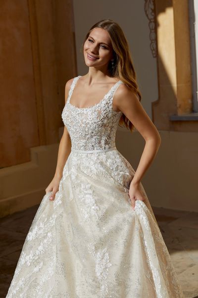 Close up of a model stood outside in Ronald Joyce 69564, a lace ballgown wedding dress with a sparkle tulle skirt, satin waist belt and straps in ivory and light gold