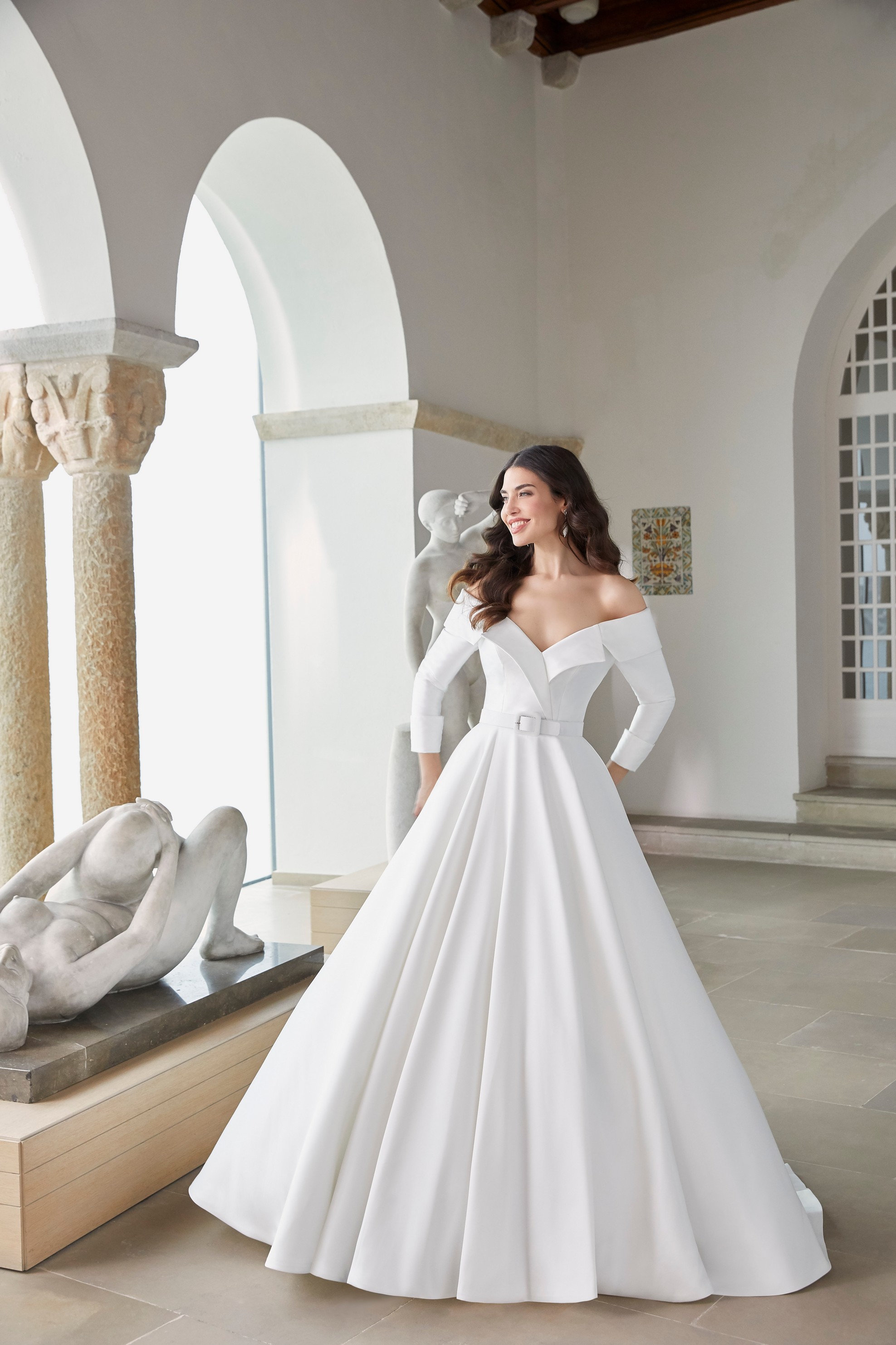 Brown haired woman standing in museum with sculptures wearing off the shoulder v neckline ballgown dress with belt