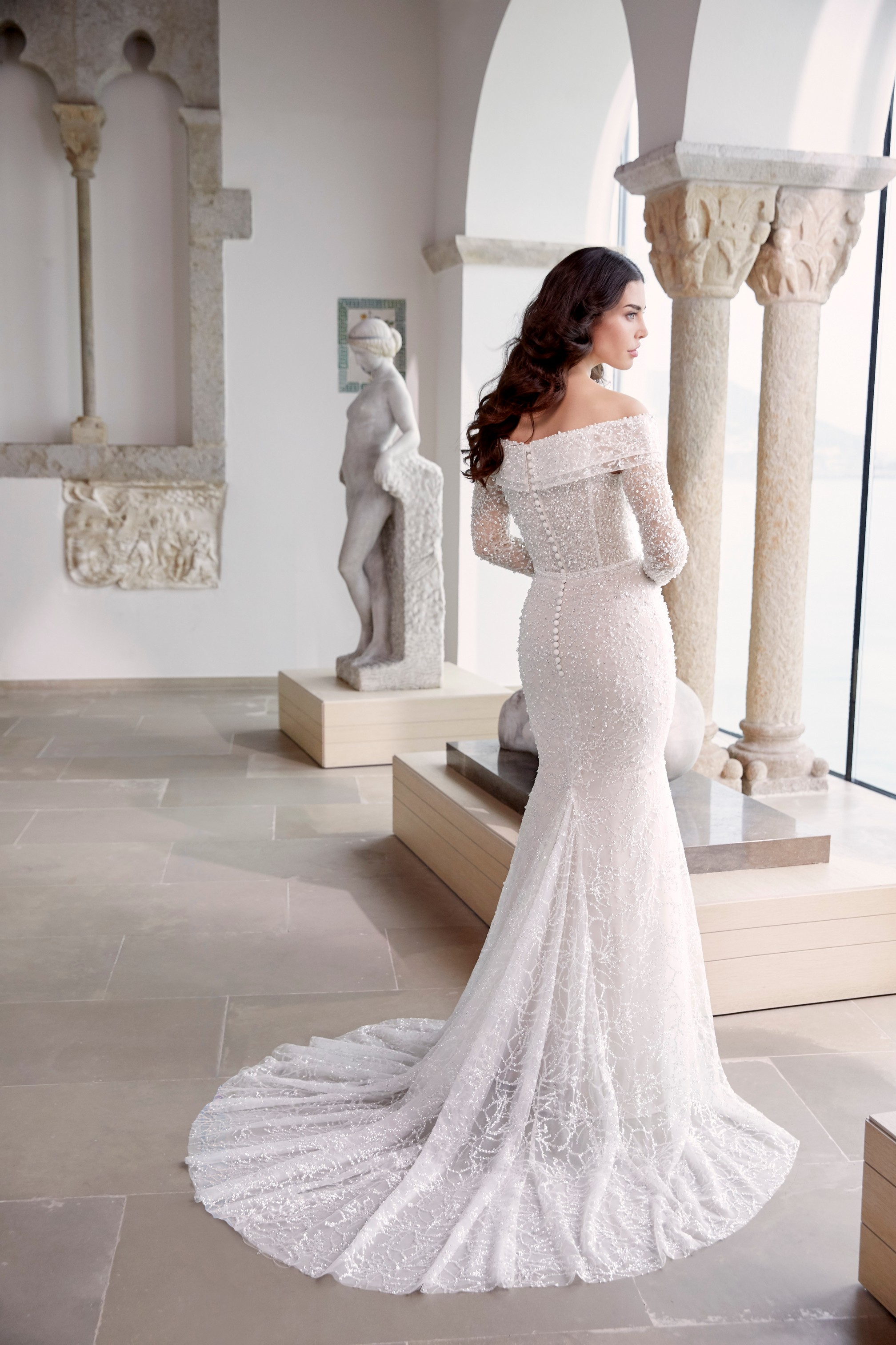 Woman stood in museum looking out of the window towards the sea wearing beaded wedding gown with off the shoulder detail 