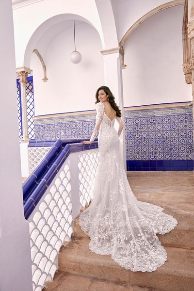 Back of model stood in a blue tiled hallway in Ronald Joyce 69667, a long sleeved wedding dress with lace appliques, sheer sleeves, a sweetheart neckline and lace scallop train