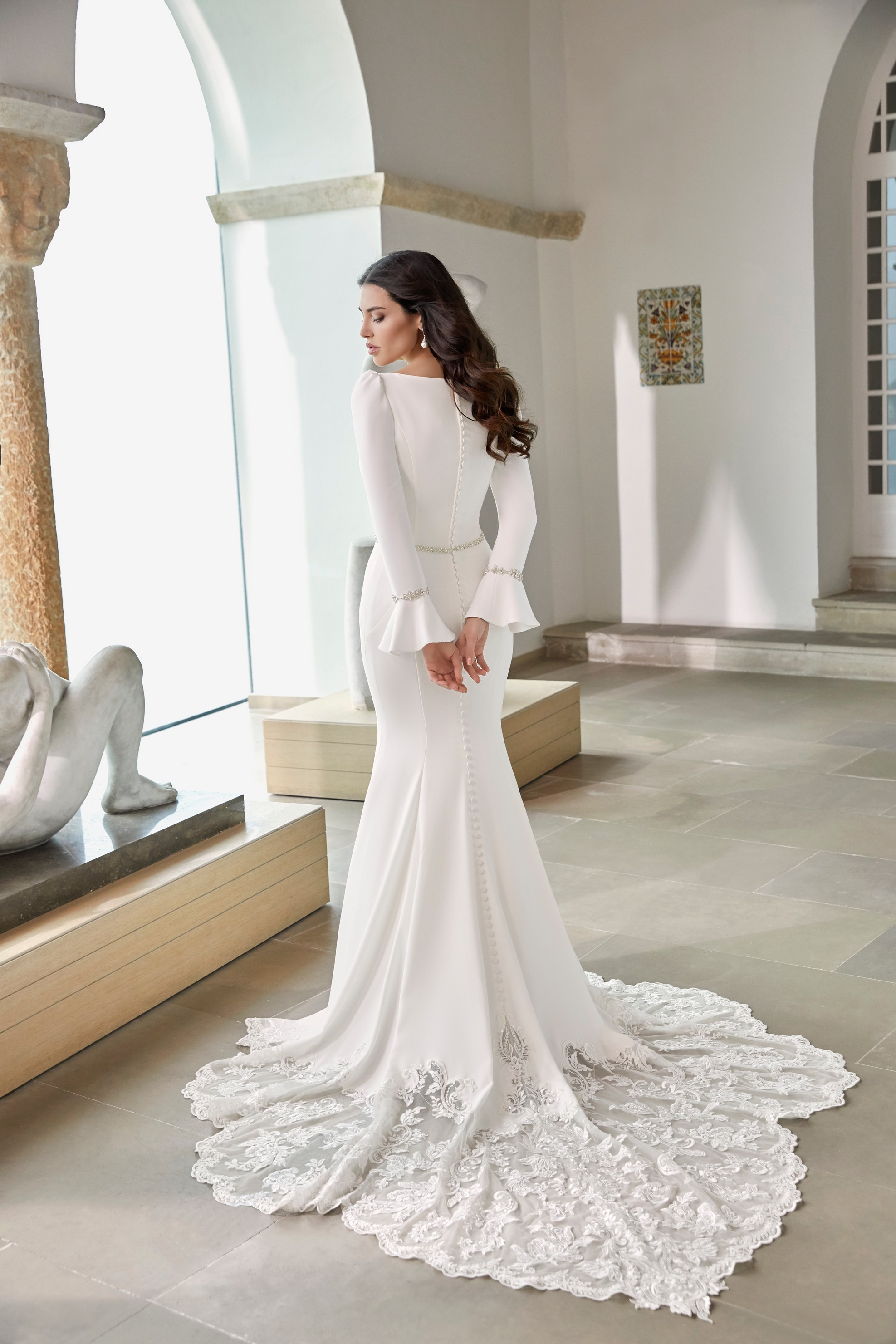 Back of model stood in an empty museum room in Ronald Joyce 69668, a long sleeved plain wedding dress with fluted cuffs, a sparkle belt, fit and flare skirt and lace scallop train