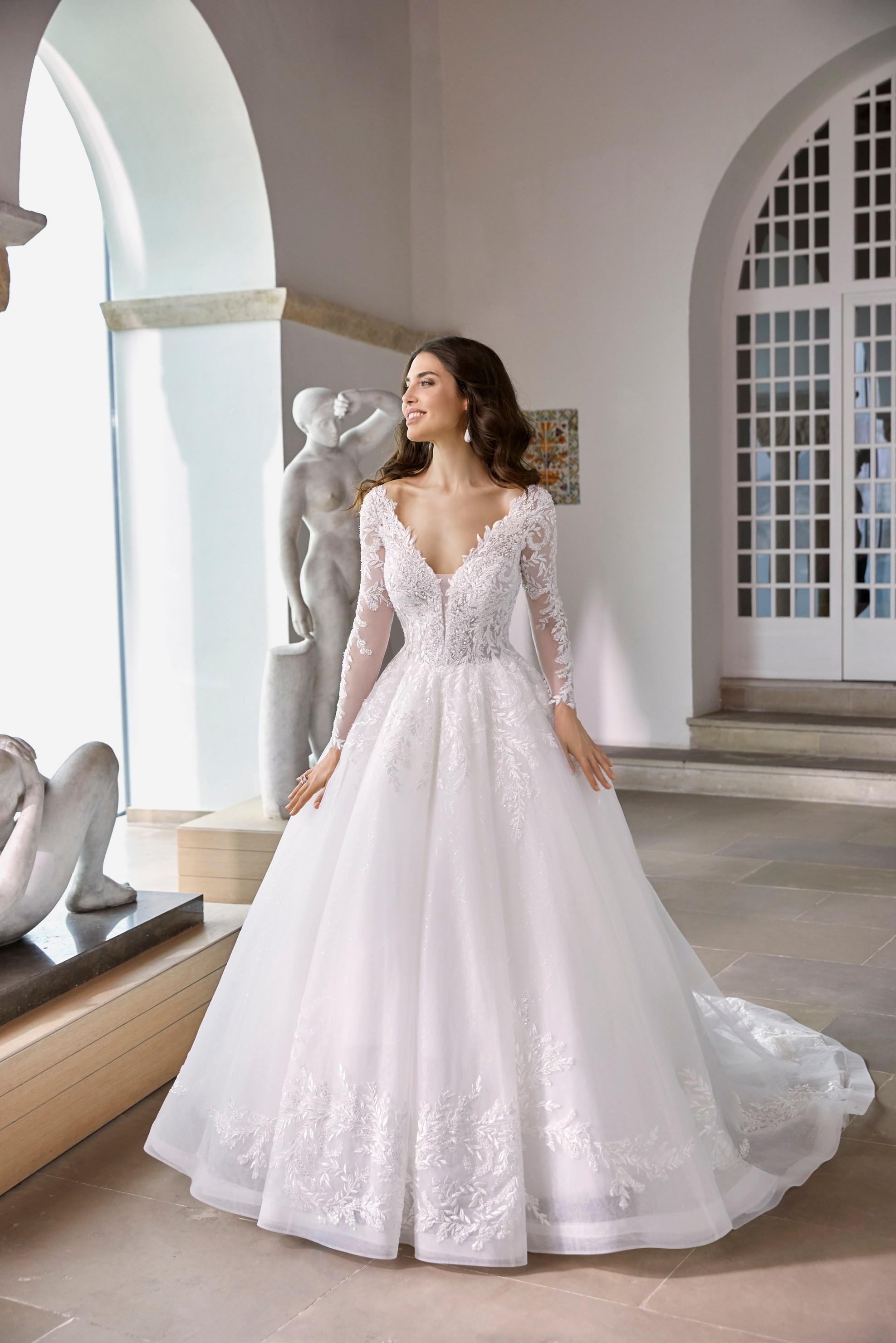 Model stood in an empty museum room in Ronald Joyce 69674, a long sleeved ballgown wedding dress with lace illusion sleeves, a plunging v-neckline and lace applique hem