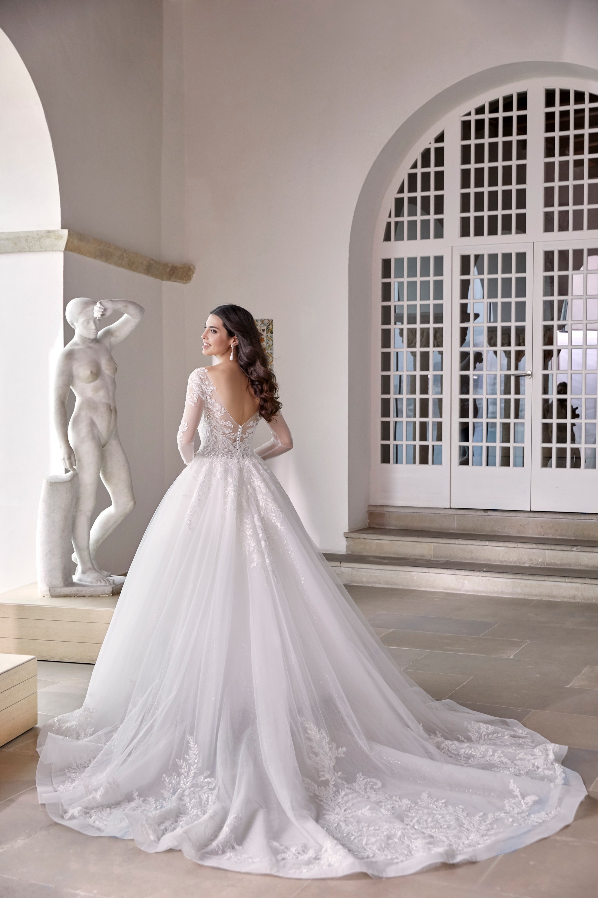 Back of model stood in an empty museum room in Ronald Joyce 69674, a long sleeved ballgown wedding dress with lace sleeves, an open back and cathedral length lace applique hem