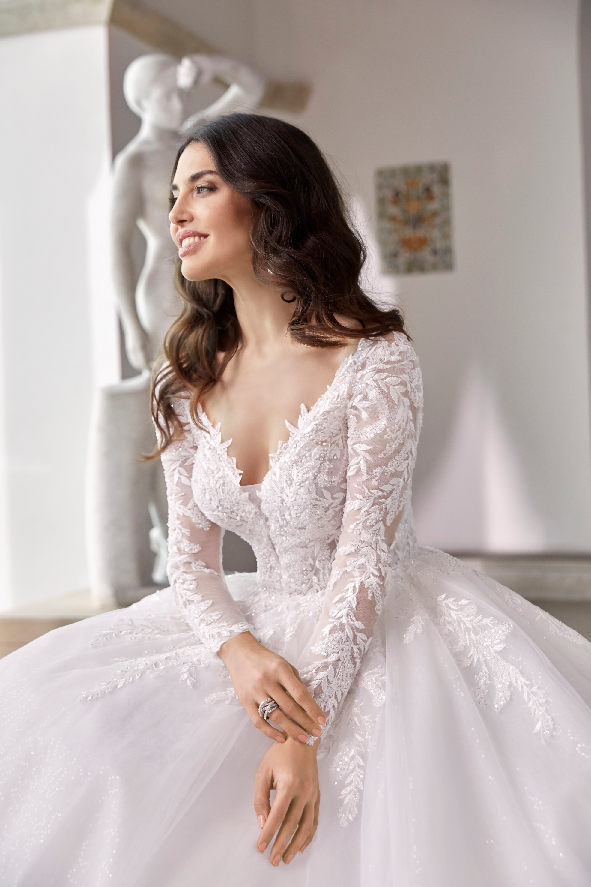 Close up of model sat in an empty museum room in Ronald Joyce 69674, a long sleeved ballgown wedding dress with lace sleeves, a v-neckline and voluminous tulle skirt