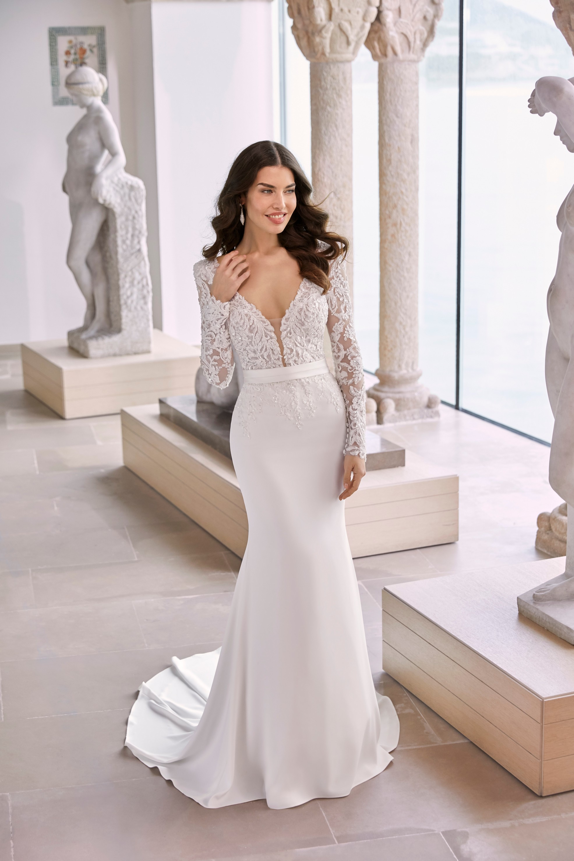 Model stood in an empty museum room in Ronald Joyce 69680, a fit and flare wedding dress with a plunging lace applique tulle bodice and matching long sleeves on a plain skirt. 