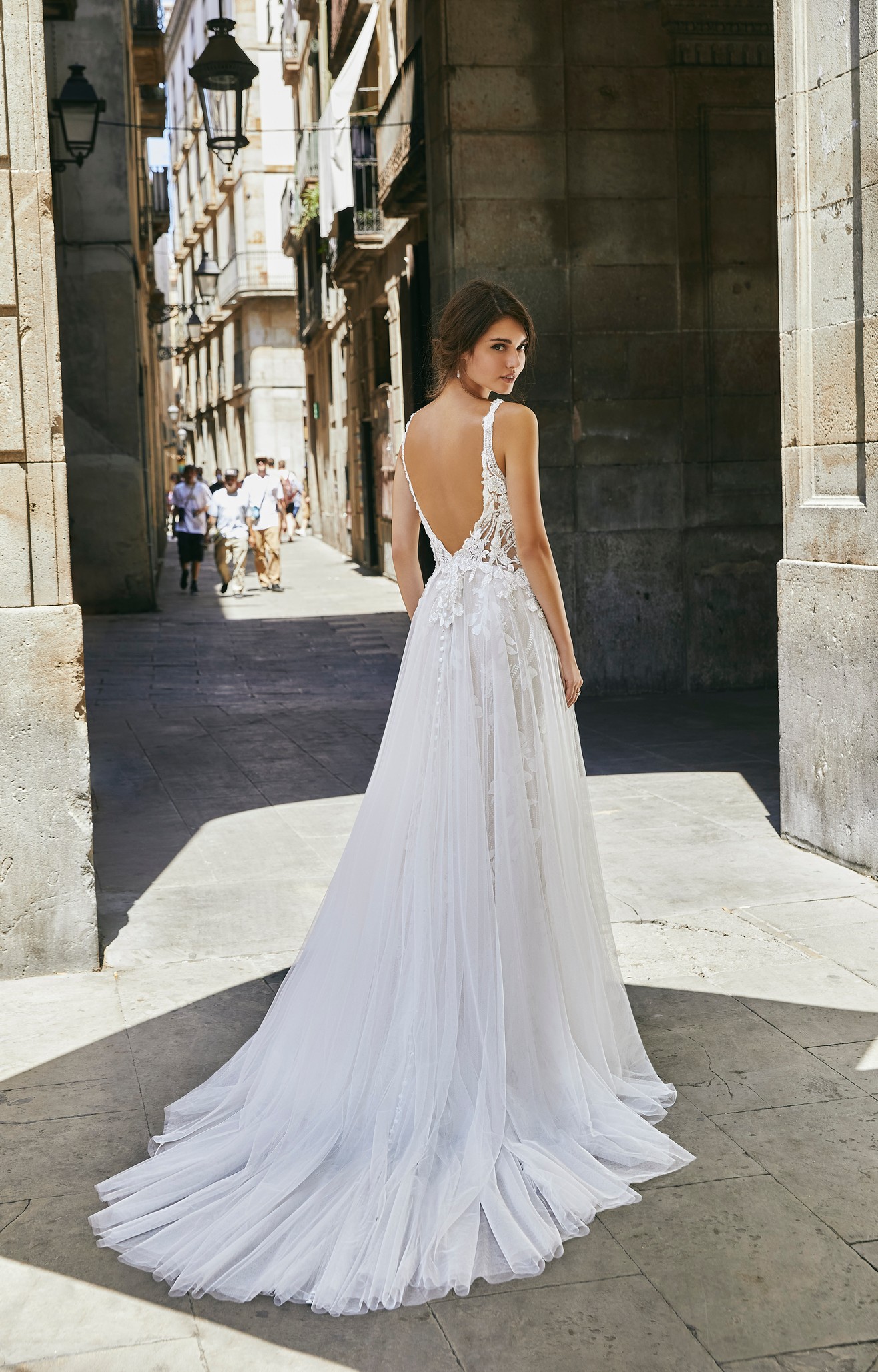 Back profile of model wearing Ronald Joyce wedding dress style 69701, a striking A-line lace wedding dress with straps, an illusion v-neckline and matching v-shape back with bridal buttons and a tulle skirt.