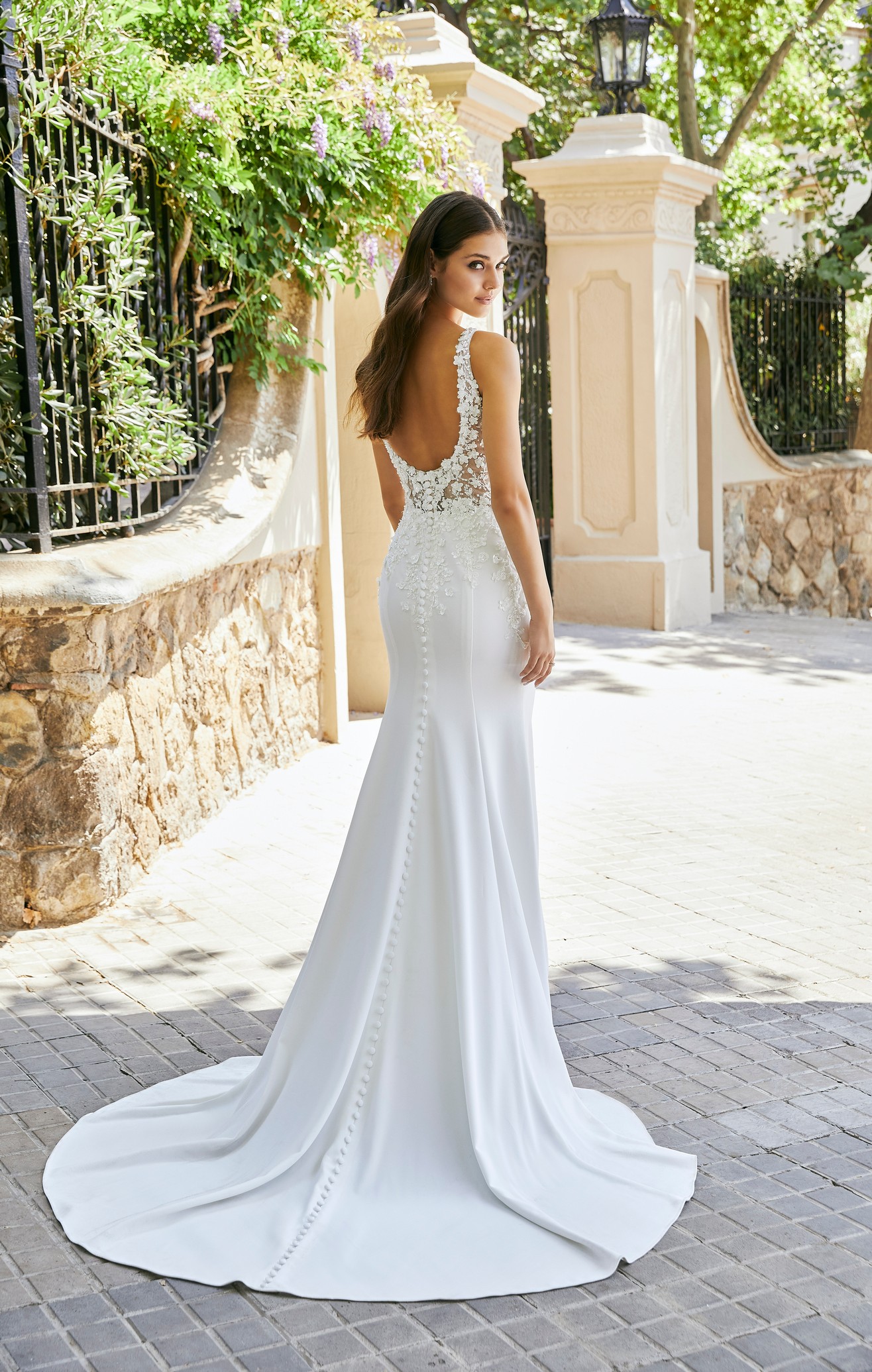 Back of model stood by a black gate in Ronald Joyce 69702, a fit and flare wedding dress with an open scoop back, 3D floral appliqué bodice and plain skirt