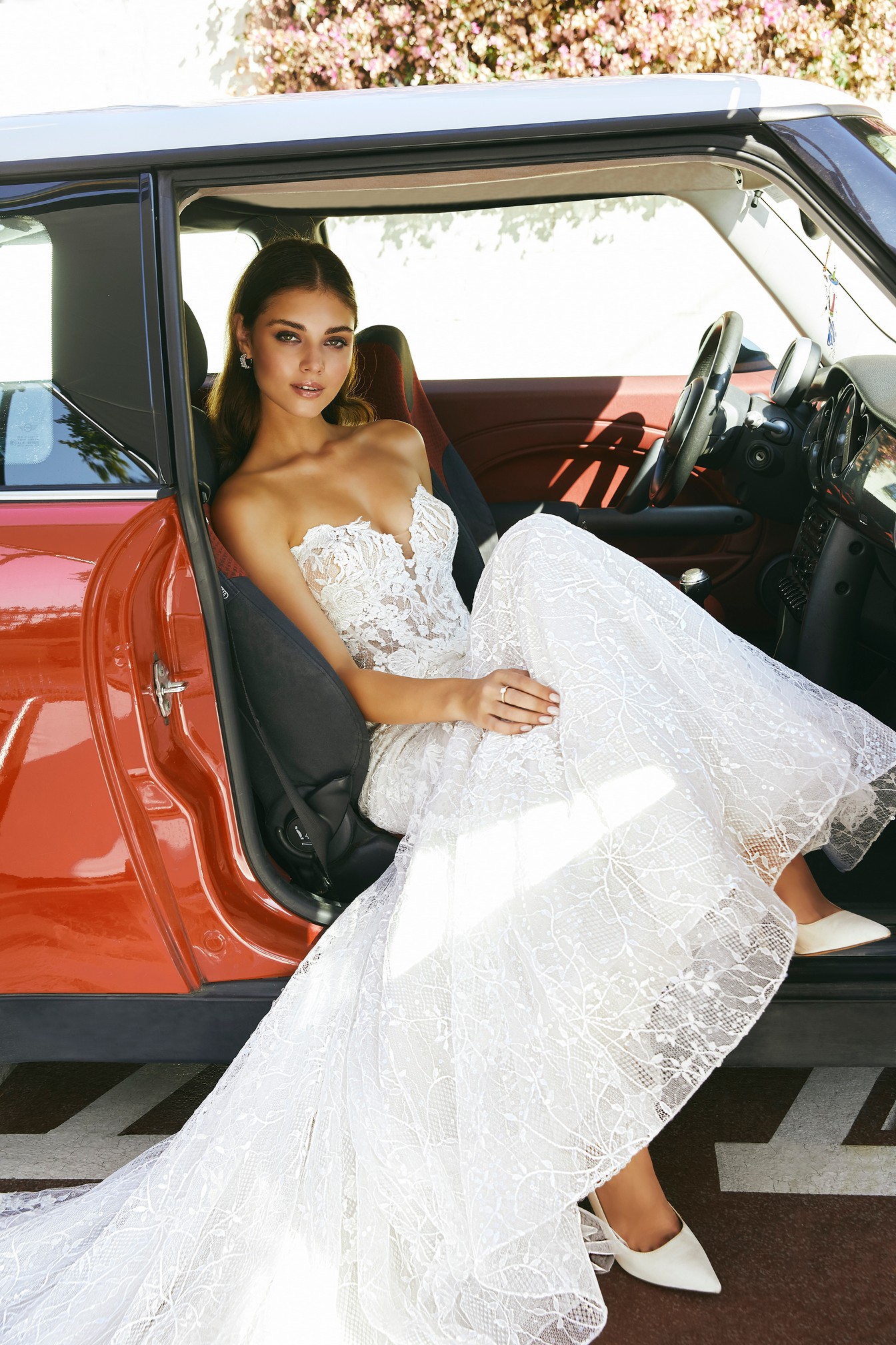 Close up of model sat in a red Mini in Ronald Joyce style 69707, a strapless lace fit and flare wedding dress with a plunging sweetheart neckline, illusion bodice and striking lace and tulle skirt 