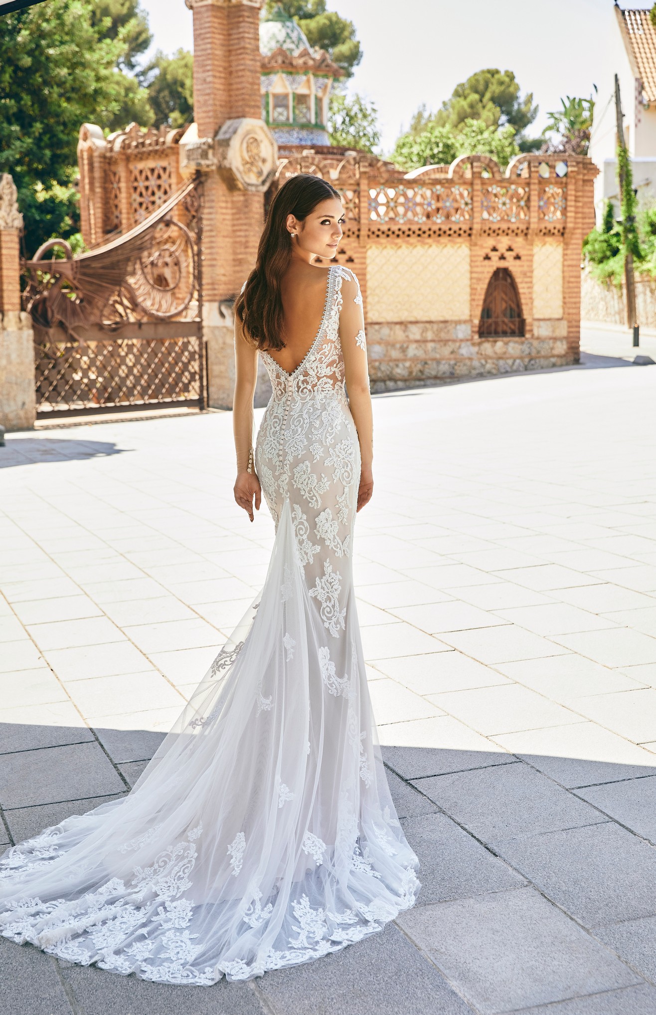 Back profile of model wearing Ronald Joyce wedding dress style 69716, a tulle and lace applique fit and flare dress with illusion full sleeves and a v-shaped back complete with button and bustle detailing. 