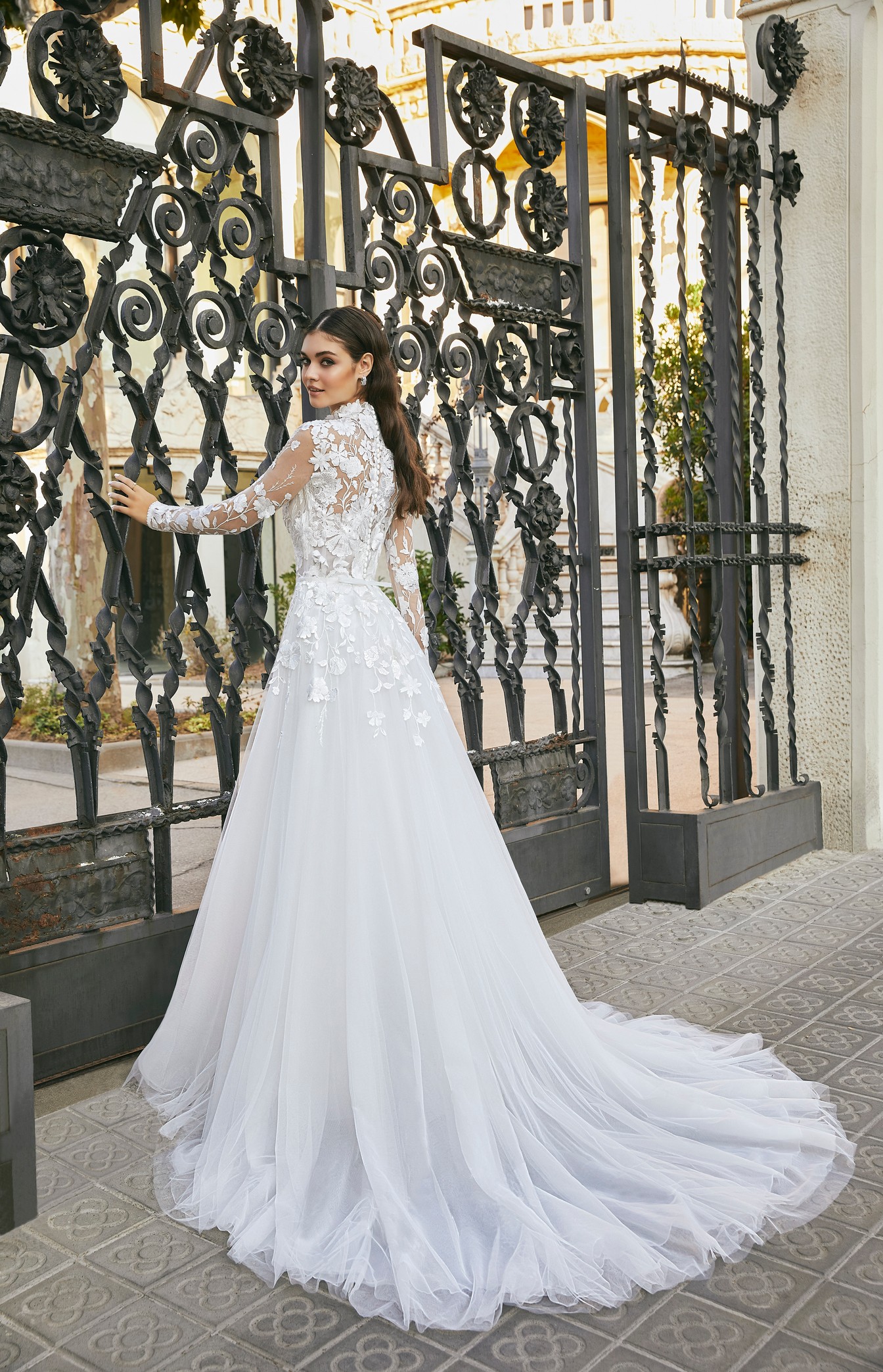 Back image of woman standing in front of decorative gates wearing ballgown wedding dress with long tulle train and lace embroidered  bodice