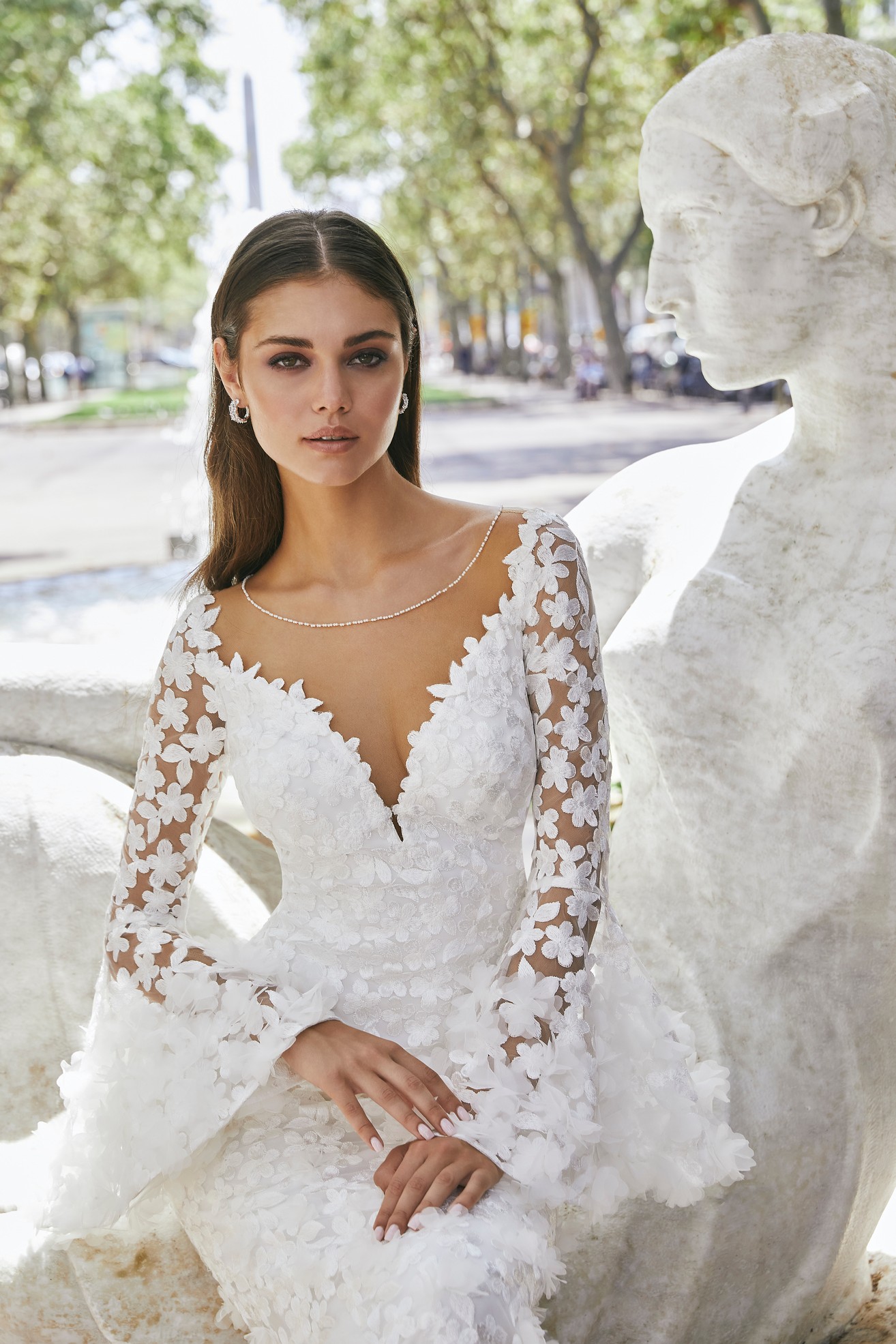 Woman sitting next to marble sculpture wearing fitted wedding dress decorated with 3-d flowers