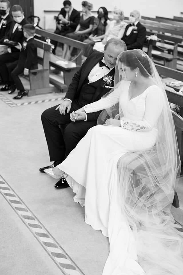 Black and white image of real bride Emma sat on a church pew next to husband Brian in her white Ronald Joyce 18202 Tai wedding dress with a floor-length tulle veil over her face.