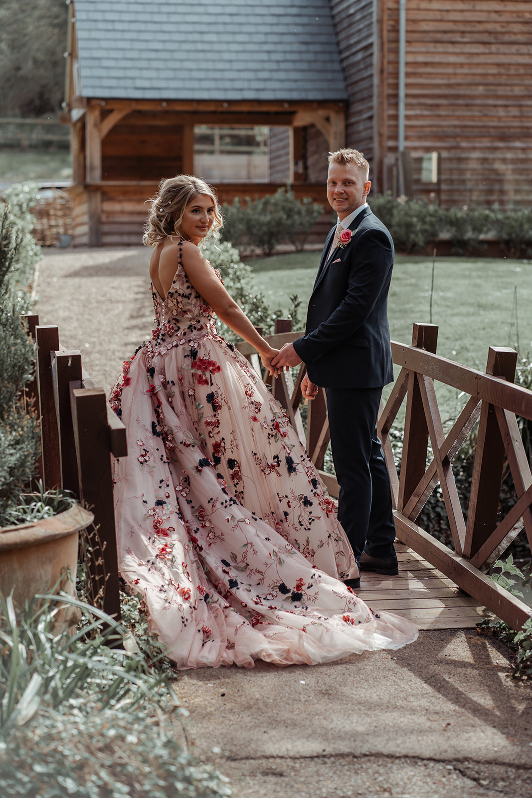 Ronald Joyce real bride, Rebecca, and her husband, Michael, holding hands on a bridge on their wedding day. Rebecca has curly blonde hair and wears Celestina,  a blush coloured ballgown wedding dress style with bold floral appliques.