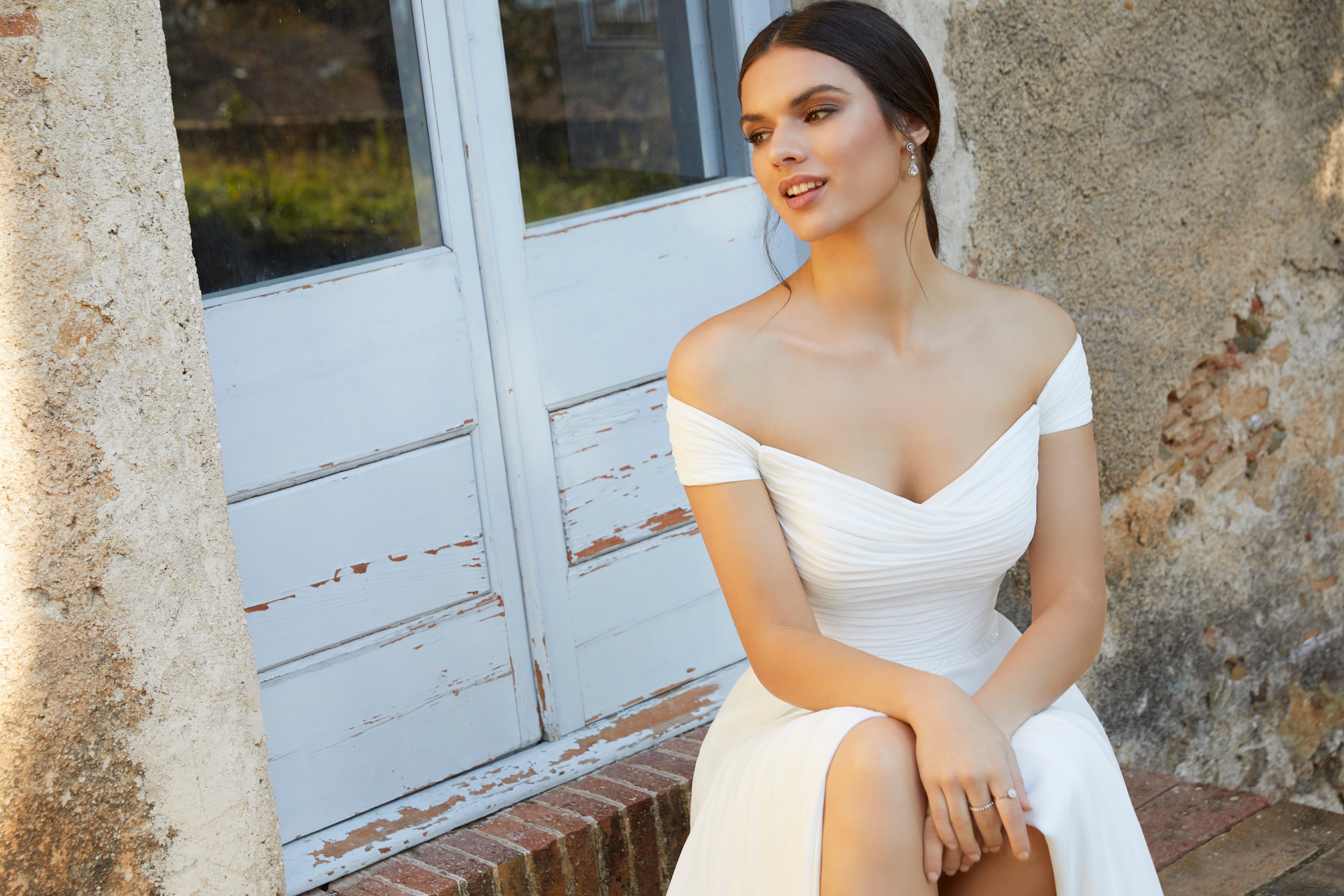 Close up of a model sat on terracotta outside steps in a bardot wedding dress (Ronald Joyce 18461), an off-the-shoulder ivory chiffon sheath dress with a ruched bodice