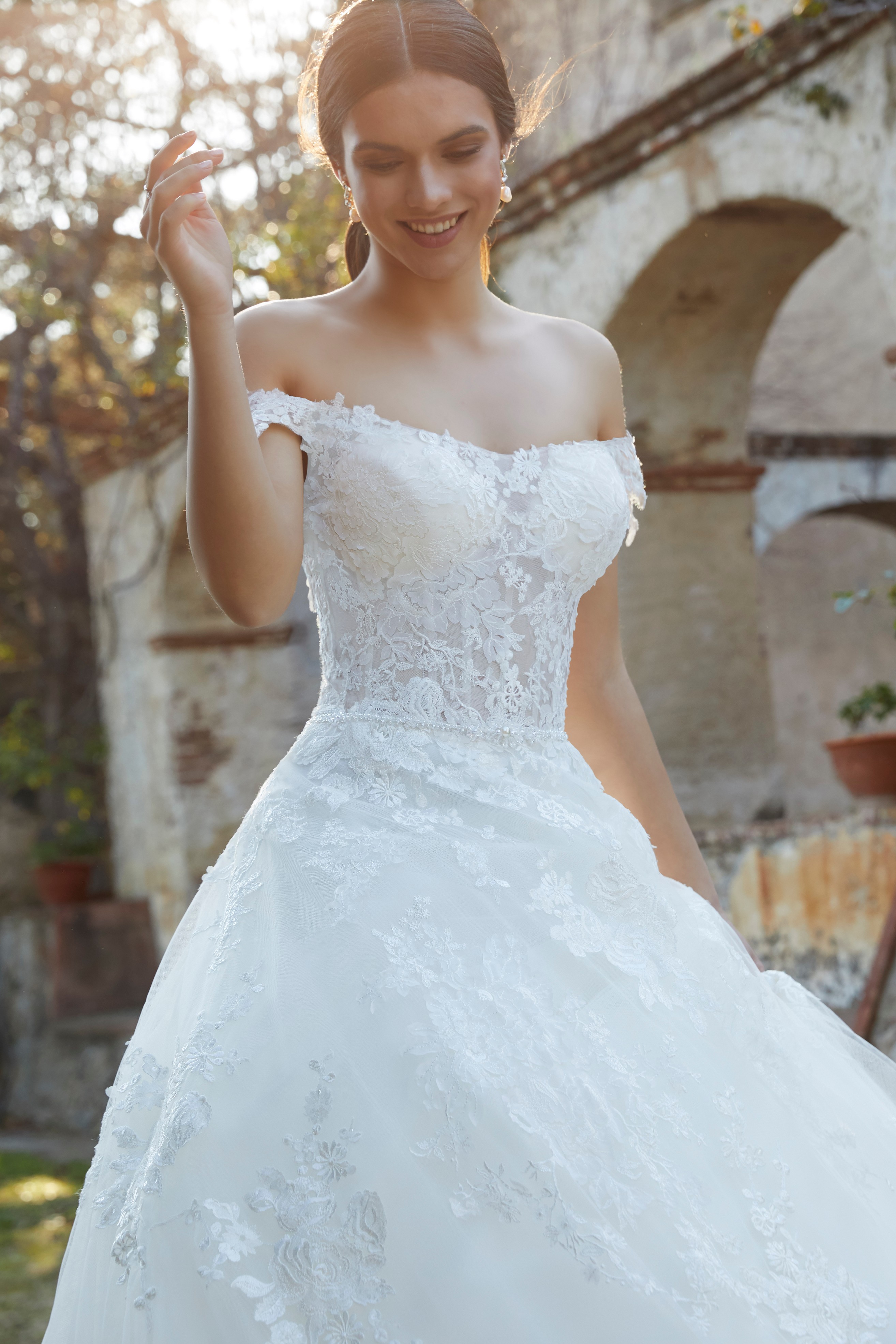 Close up of a model stood outside in a bardot wedding dress (Ronald Joyce 18470), an off-the-lace ballgown dress with a sheer illusion bodice and lace applique skirt