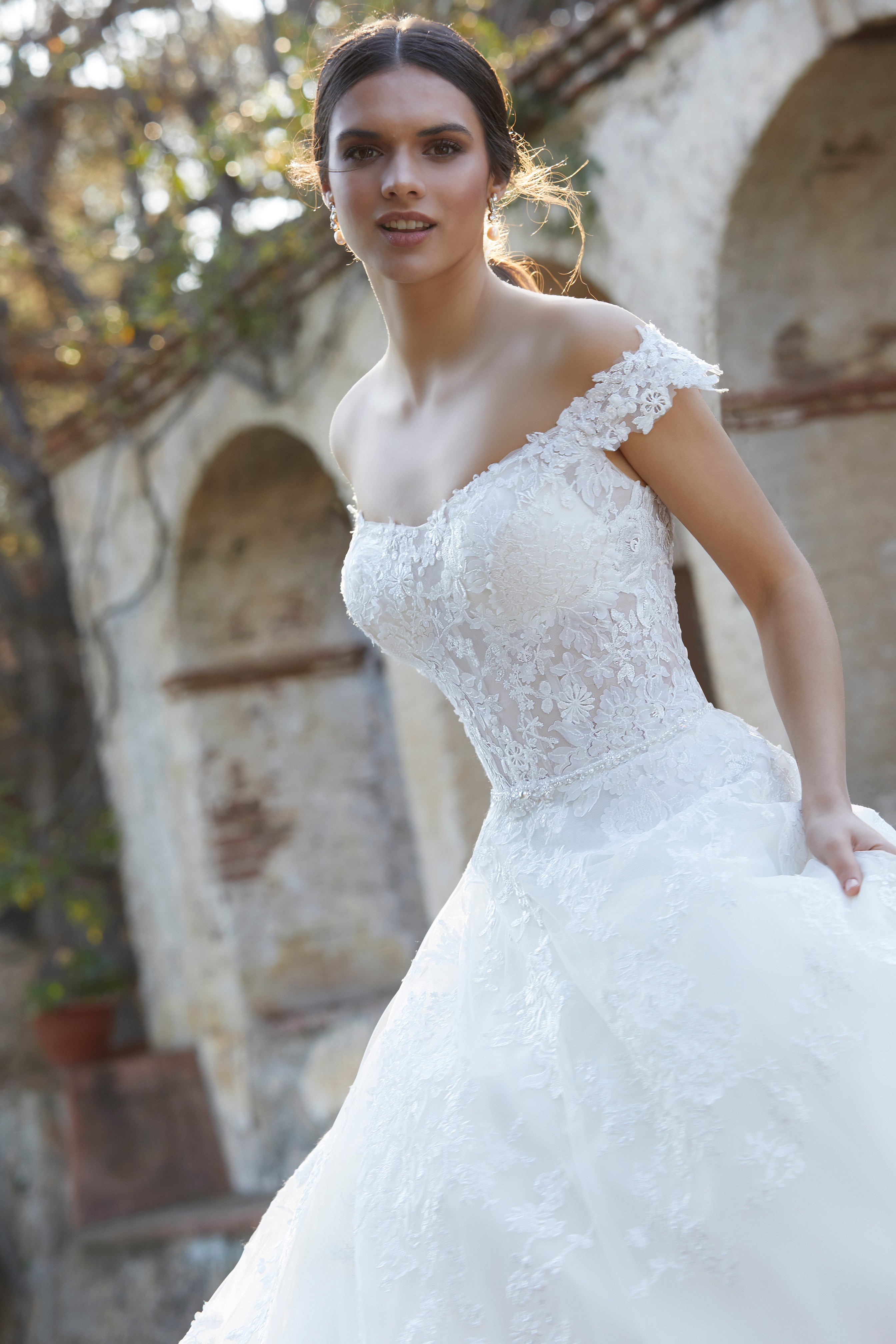 Thumbnail photo of model stood outside by archways in Ronald Joyce 18470, an ivory lace A-line wedding dress with off-the-shoulder bardot-style sleeves